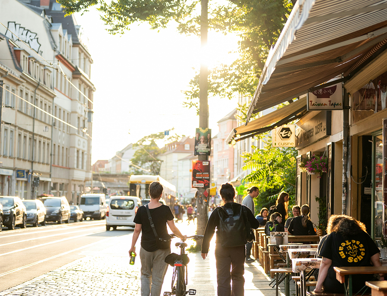 Straßenszene mit Menschen, Café und Fahrrädern bei Sonnenuntergang.